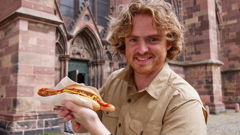 Nomadic Samuel enjoying a freshly grilled Lange Rote sausage in front of the Gothic Freiburg Cathedral in Freiburg im Breisgau, Germany — a must-try local specialty during a visit to the city