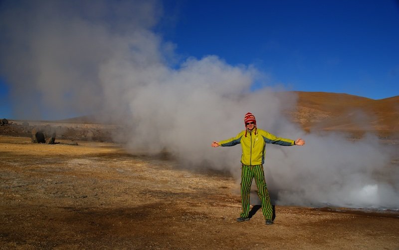 Nomadic Samuel standing with open arms in front of steaming geothermal vents at El Tatio Geysers, Atacama Desert, Chile