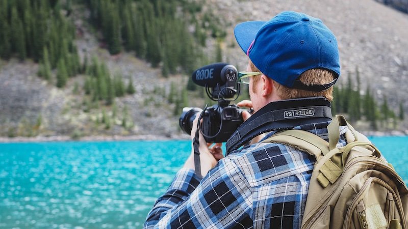 Nomadic Samuel filming with a camera beside the vivid turquoise waters of Moraine Lake, capturing scenic views of the surrounding forested slopes and rugged peaks during our Canadian Rockies road trip