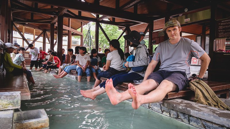 Nomadic Samuel relaxing with his feet soaking in the free public foot bath at Yumoto Onsen in Nikko, Japan, surrounded by locals and visitors enjoying the warm mineral waters under a wooden pavilion