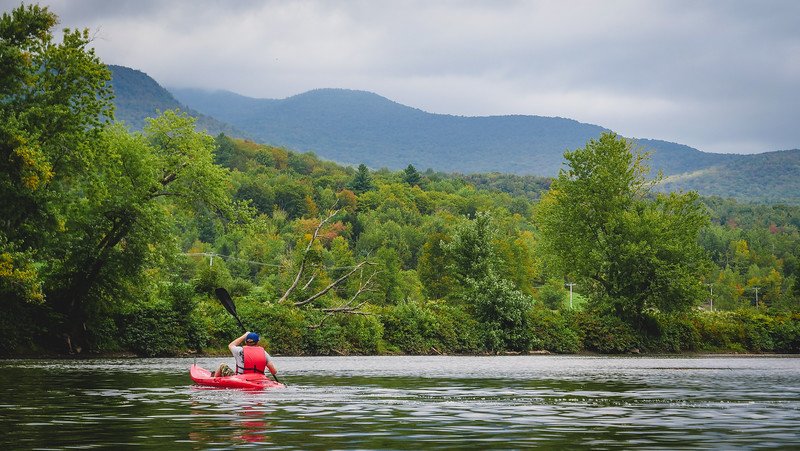 Nomadic Samuel kayaking on the serene Missisquoi River surrounded by forested hills and the rolling mountains of the Eastern Townships near Sutton, Quebec, enjoying a peaceful outdoor adventure