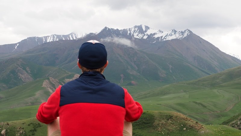 Nomadic Samuel sitting on a hillside in a red and navy jacket, admiring the sweeping green valleys and snow-capped mountain peaks of the Tian Shan range during the 3-Day Panorama Trek in Kyrgyzstan