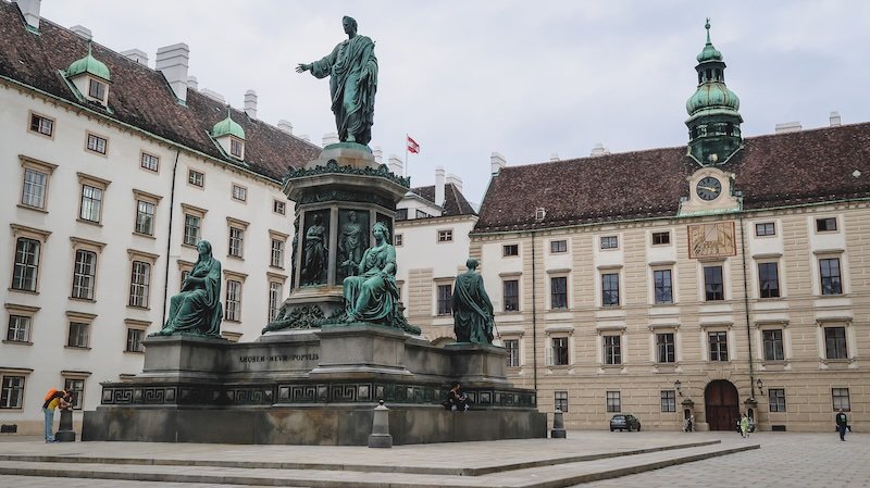 Nomadic Samuel taking a photo from the far left corner while capturing the elegant historic architecture of Vienna, Austria, with grand buildings, ornate façades, and bustling pedestrians in the city center Nomadic Samuel taking a photo from the far left corner while capturing the elegant historic architecture of Vienna, Austria, with grand buildings, ornate façades, and bustling pedestrians in the city center