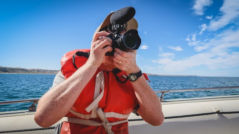 Nomadic Samuel wearing a bright orange life jacket captures the scenery with his camera during a boat tour in Peninsula Valdés, Patagonia, Argentina, a UNESCO World Heritage Site known for wildlife, dramatic coastlines, and eco-adventure travel