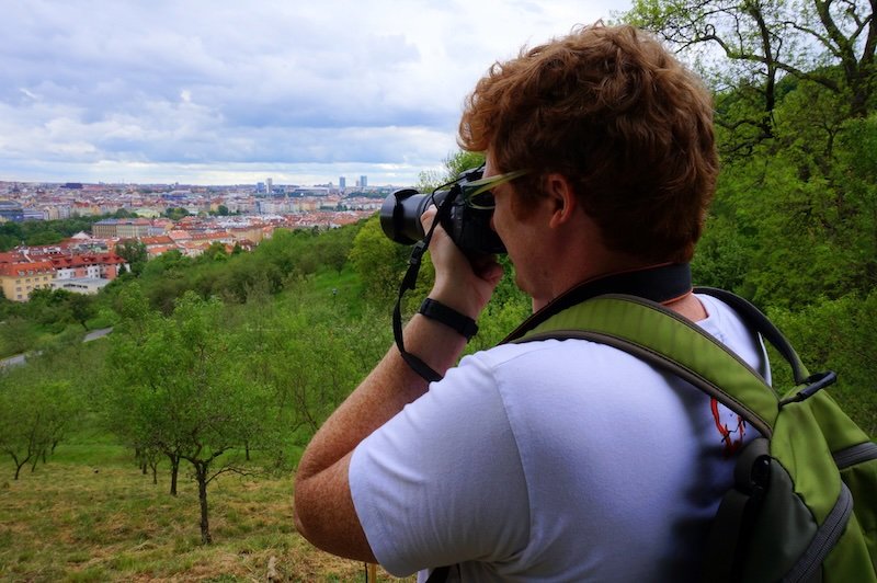 Nomadic Samuel taking photographs from a scenic hilltop viewpoint overlooking the red rooftops and cityscape of Prague, Czech Republic, surrounded by lush greenery