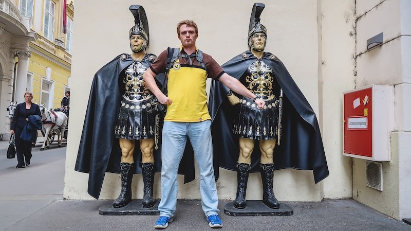 Nomadic Samuel striking a humorous pose between two ornate Roman-style guard statues in Vienna, Austria, blending in playfully with the figures Nomadic Samuel striking a humorous pose between two ornate Roman-style guard statues in Vienna, Austria, blending in playfully with the figures