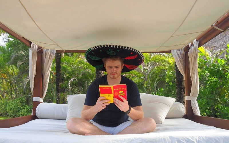 Nomadic Samuel relaxing on a shaded daybed at Royal Hideaway Playacar in Playa del Carmen, wearing a colorful sombrero and reading a travel book amidst lush tropical gardens