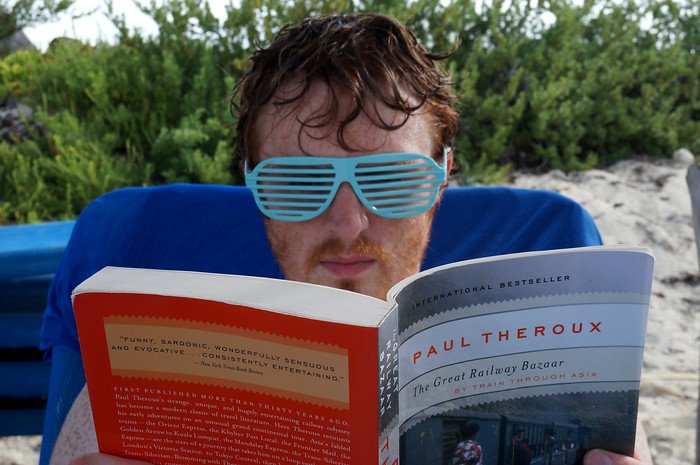 Nomadic Samuel relaxes on a beach chair wearing playful blue shutter shades while reading The Great Railway Bazaar by Paul Theroux, enjoying a laid-back afternoon on a tropical beach in Cayo Santa María, Cuba