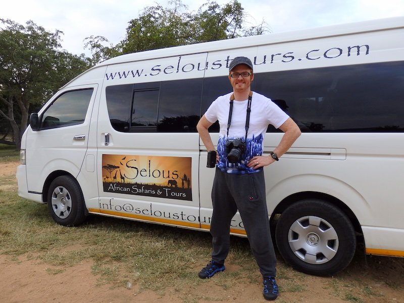 Nomadic Samuel dressed in a casual safari outfit with a cap, t-shirt, and comfortable pants, standing next to a Kruger National Park safari tour van with his camera ready for wildlife photography in South Africa