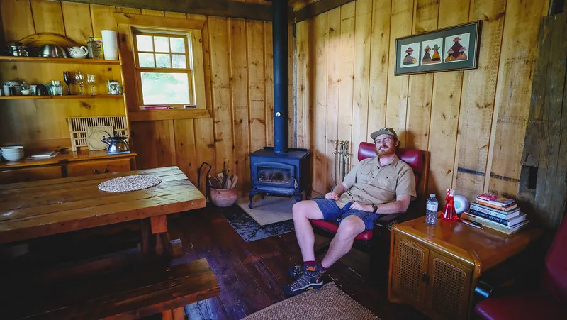 Nomadic Samuel sitting inside the off-grid cabin with an all wood interior plus a wood-burning stove Nomadic Samuel sitting inside the off-grid cabin with an all wood interior plus a wood-burning stove
