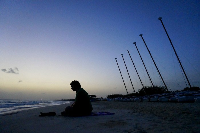 In the soft pre-dawn light, Nomadic Samuel sits in peaceful silhouette on the sandy shore as the sun rises over the calm turquoise waters of Cayo Santa María, Cuba, with sailboats lined up along the beach nearby