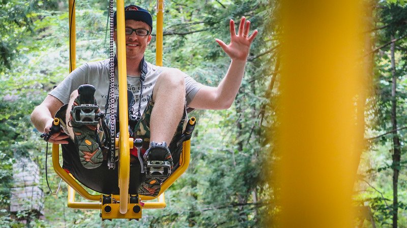 Nomadic Samuel smiling and waving while pedalling the bright yellow Vélo Volant canopy bicycle through the treetops at Au Diable Vert in Sutton, Quebec, enjoying an adventurous ride in the Eastern Townships