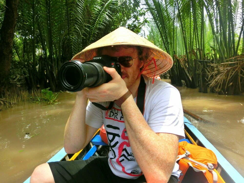 Nomadic Samuel photographing from a boat in Vietnam’s Mekong Delta wearing a traditional conical hat, capturing river life with a Sony DSLR