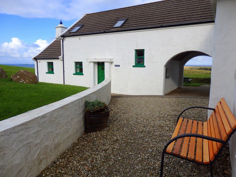 Charming whitewashed cottage with green trim nestled along the rugged coastline of Northern Ireland, featuring a gravel courtyard with a wooden bench, arched passageway, and sweeping views of the surrounding countryside and sea under a bright blue sky