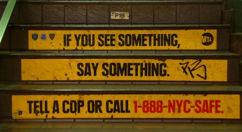 Yellow safety signs on the steps of a New York City subway station display the iconic “If You See Something, Say Something” message with the MTA logo and emergency number 1-888-NYC-SAFE, reminding riders of the city’s public safety campaign