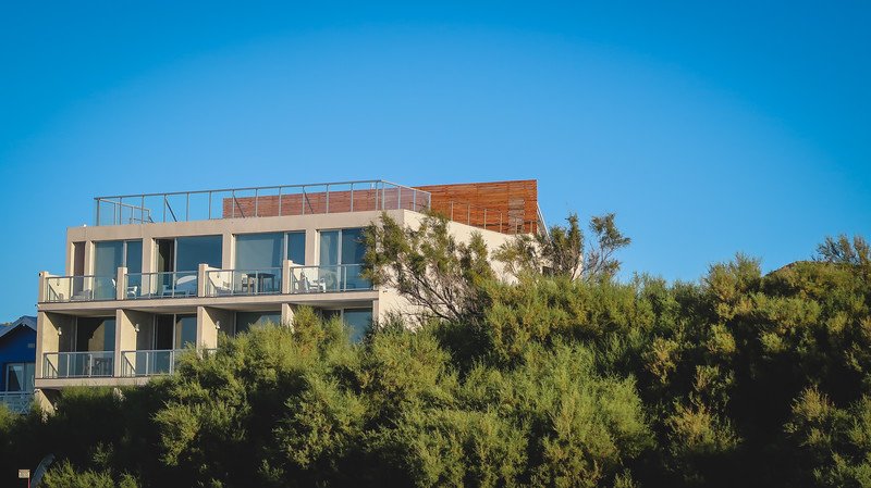 Modern eco-friendly Oceano Patagonia hotel in Puerto Pirámides, Península Valdés, Argentina, framed by lush native vegetation under a clear blue sky. The minimalist design with balconies and a rooftop terrace reflects sustainable coastal architecture.