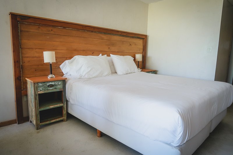 Bright and minimalist hotel room at Oceano Patagonia in Puerto Pirámides, Península Valdés, Argentina, featuring a comfortable king-size bed with crisp white linens, rustic wooden headboard, and vintage bedside tables — a cozy eco-lodge retreat