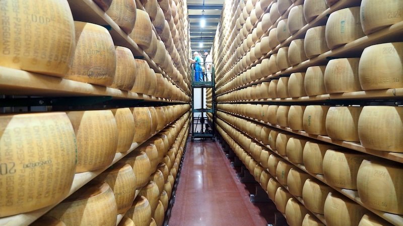 Endless rows of Parmigiano-Reggiano cheese wheels aging on tall wooden shelves inside a dairy in Emilia-Romagna, Italy, with workers inspecting the prized cheeses, showcasing the region’s artisanal food traditions and world-renowned culinary heritage