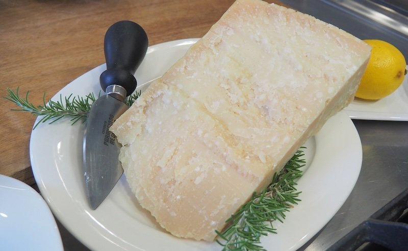 Large wedge of aged Parmigiano-Reggiano cheese served on a white plate with a traditional knife, garnished with rosemary and lemon, highlighting Emilia-Romagna’s world-famous culinary heritage and the authentic flavors of Italian food culture