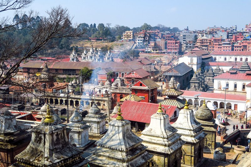 Smoke rising from Hindu cremation ghats along the Bagmati River at Pashupatinath Temple in Kathmandu, Nepal, with sacred shrines, courtyards, and cityscape in the background