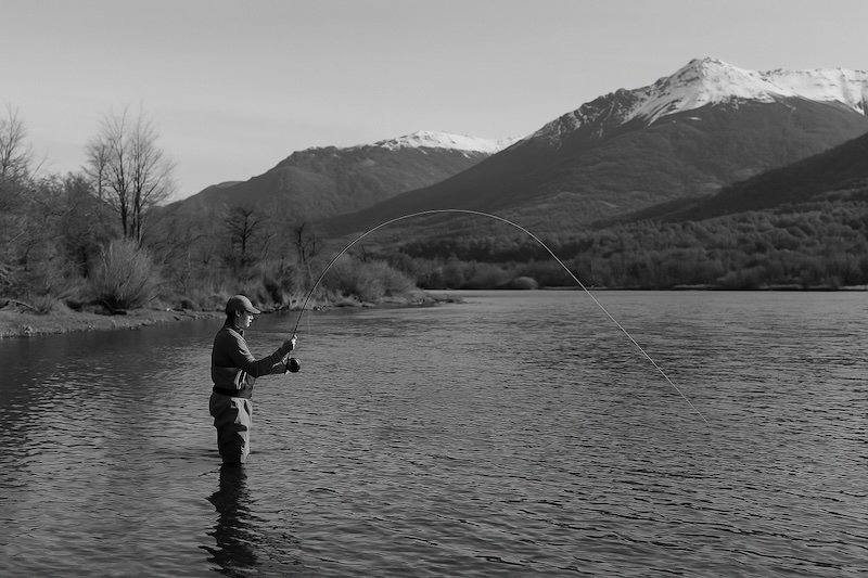 A black and white photograph of a fly fisherman standing mid-river in Patagonia, Argentina. Wearing waders, he casts a light 3-weight fly rod over calm water. Bare autumn trees and snow-dusted mountains form a crisp, clear backdrop under an open sky