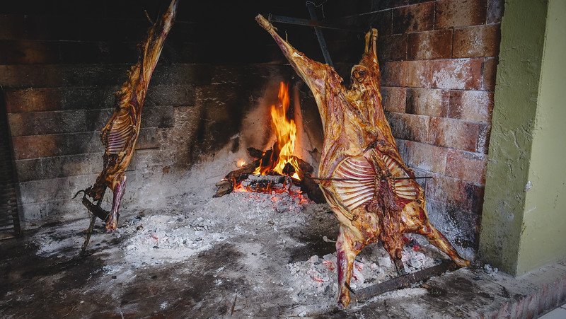 Traditional Patagonian lamb asado cooking on an open fire at Estancia San Lorenzo on Península Valdés, Argentina. Two whole lambs are stretched on iron crosses and slow-roasted beside glowing embers inside a rustic brick parrilla, preparing a classic countryside lunch