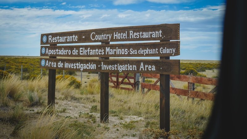 Directional wooden signpost at Península Valdés in Patagonia, Argentina, indicating the way to a country hotel restaurant, a sea elephant colony viewing area, and an investigation zone. The sign stands in a windswept steppe landscape under a cloudy sky