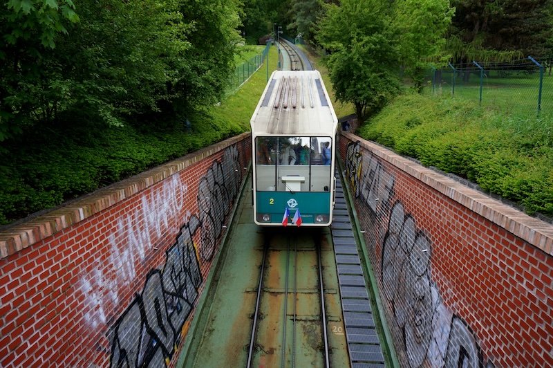 Green funicular car traveling up the historic Petřín Funicular railway through a tree-lined slope and graffiti-covered brick walls in Prague, Czech Republic