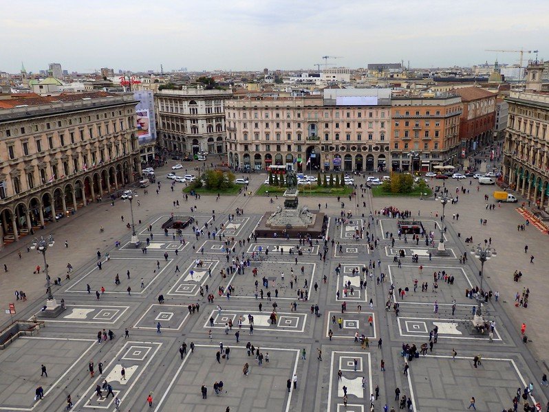Aerial view of the grand Piazza del Duomo in Milan, Italy, showing the elegant neoclassical buildings, geometric square patterns, and bustling crowds seen from the rooftop of Duomo di Milano