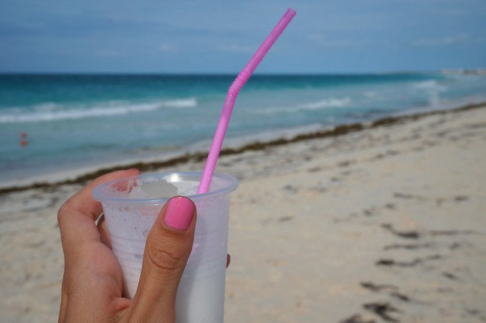 A hand with pink nail polish holds a chilled piña colada in a clear plastic cup with a pink straw, with turquoise waves and soft white sand in the background on a sunny beach in Cayo Santa María, Cuba