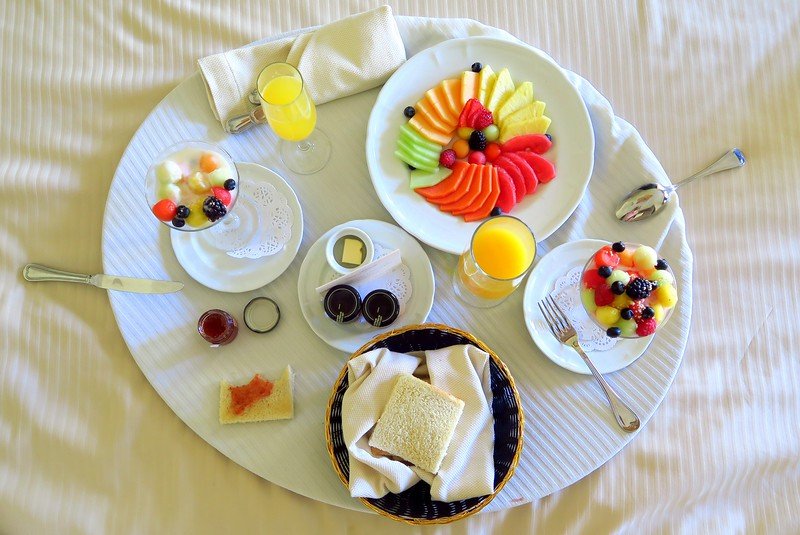 Colorful tropical breakfast spread served in-room at a Playa del Carmen hotel, featuring a vibrant platter of sliced tropical fruits, fresh fruit parfaits, toast, orange juice, and jam, artfully arranged on a white round tray atop crisp linens