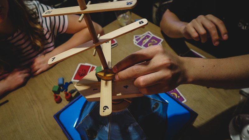 Close-up of friends playing a balancing board game at Draughts café in London, with hands carefully placing wooden pieces on a tower, colorful cards on the table, and a lively atmosphere that captures the social fun of board gaming in the city