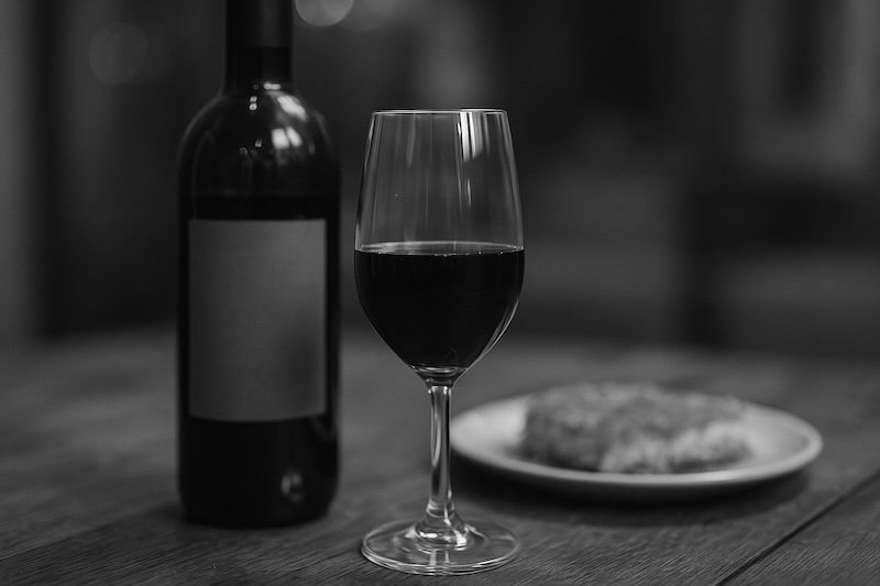 A black and white photograph of a bottle and glass of pomegranate wine from the Galilee, placed on a rustic wooden table. The bottle’s label is softly blurred, while the filled glass is in focus. A plate of knaffe dessert is blurred in the background, creating an elegant pairing scene