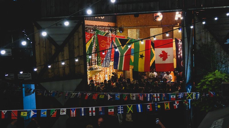 Colorful international flags, including South Africa, Ghana, and Canada, hanging above a lively crowd at Pop Brixton in London, with twinkling string lights and festive decorations creating a vibrant multicultural atmosphere at this community hub