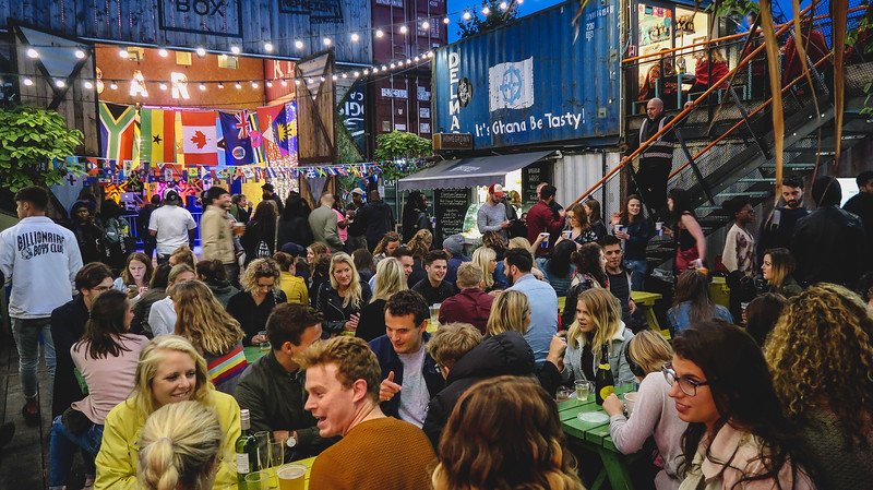 Bustling evening atmosphere at Pop Brixton in London with crowds enjoying food, drinks, and conversation under string lights, colorful flags, and repurposed shipping container stalls, showcasing the city’s vibrant street food and nightlife culture