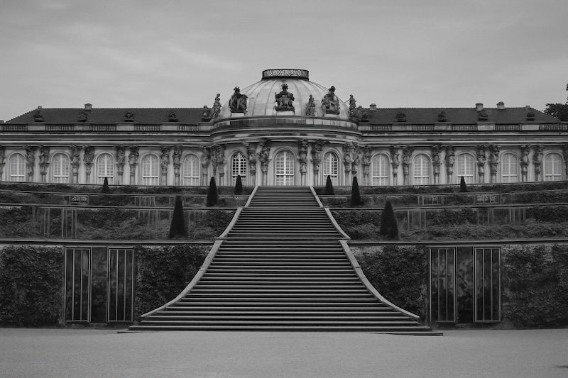 A black and white photograph of Sanssouci Palace in Potsdam, Germany, captured from a low vantage point with straight, symmetrical stairs leading up through terraced gardens to the Rococo palace. The ornate dome, statues, and columns are sharply defined against a cloudy sky