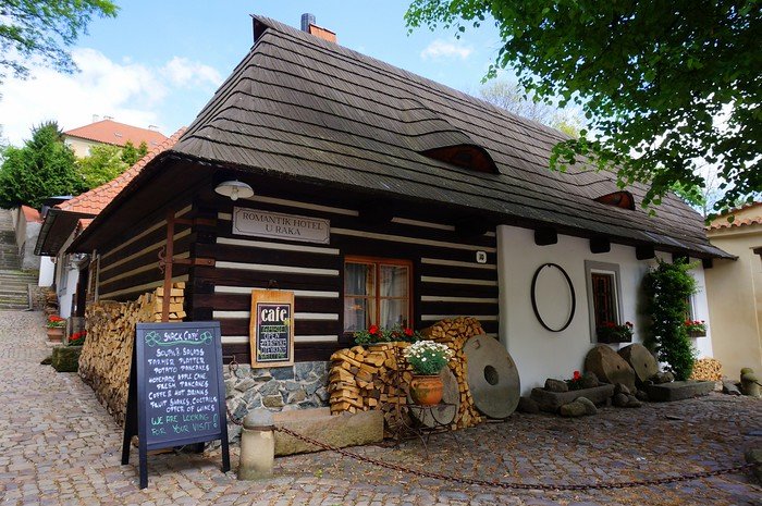A charming timber-framed cottage café in Prague with a cobblestone courtyard, stacked firewood, and blooming flowers, showcasing the city’s rustic architecture and cozy atmosphere in one of Europe’s most picturesque capitals