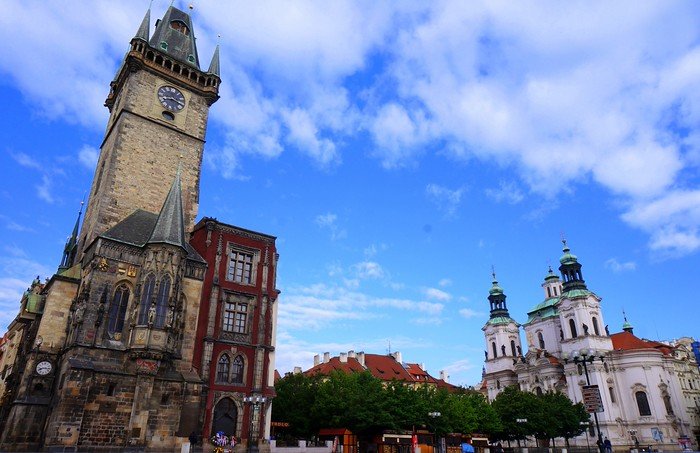 Prague’s Old Town Square featuring the Gothic Old Town Hall tower with its clock on the left and the baroque St. Nicholas Church on the right, surrounded by red-roofed buildings and a lively square under a bright blue sky with scattered clouds