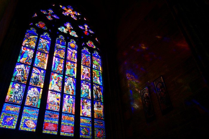 Stained glass window in Prague, Czech Republic, with vibrant colors and detailed biblical scenes illuminated by sunlight, casting colorful reflections on the stone walls of the church interior, highlighting the city’s rich Gothic artistry