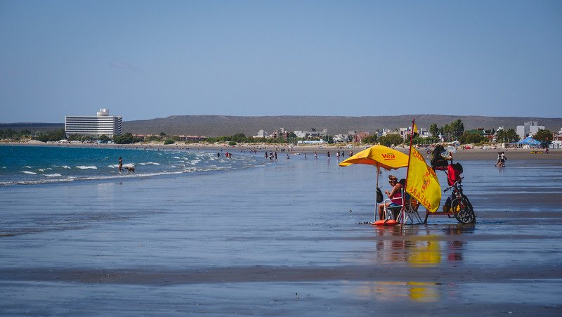 Visitors relax under a bright yellow umbrella on the wide sandy beach in Puerto Madryn, Argentina, with gentle waves rolling in and the town’s modern buildings visible in the distance, a favorite spot for sun and sea lovers