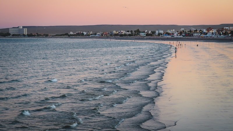 Sunset paints soft pastel hues across the sky and shoreline in Puerto Madryn, Patagonia, Argentina, where gentle waves meet the sandy beach and the coastal city skyline stretches along the horizon, offering a tranquil view of the Atlantic Ocean