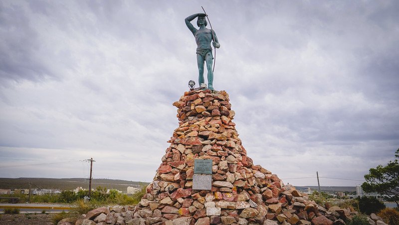 A striking monument along the Puerto Madryn waterfront in Patagonia, Argentina, featuring a statue of a Tehuelche indigenous hunter with bow and arrow, standing tall atop a pyramid of stones, symbolizing the region’s cultural history and native heritage