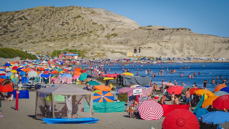 Colorful umbrellas and beach tents line the sandy shore of Puerto Pirámides on Península Valdés, Patagonia, Argentina, as crowds of people enjoy a sunny summer day swimming and relaxing by the calm blue waters surrounded by desert cliffs