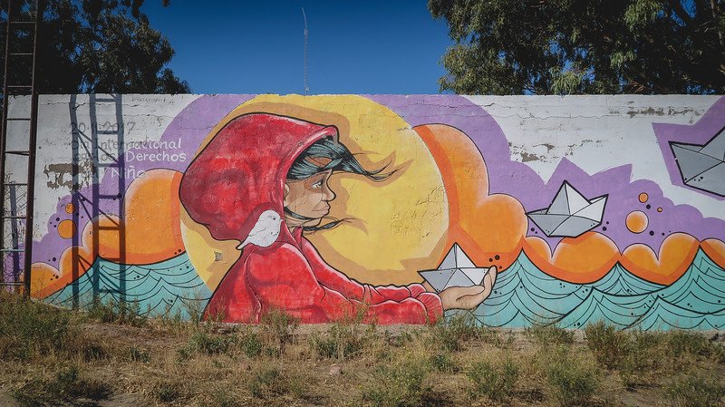 Colorful street art mural in Puerto Pirámides, Patagonia, depicting a child in a red hood releasing paper boats into stylized waves beneath a vivid sunset sky. This vibrant artwork highlights the town’s creative spirit and cultural expression through murals