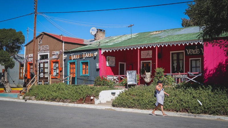 Colorful storefronts and rustic buildings line the main street of Puerto Pirámides, a charming beachside town on Península Valdés, Argentina. The vibrant facades, handmade signs, and casual pedestrian scene capture the relaxed coastal vibe of Patagonia