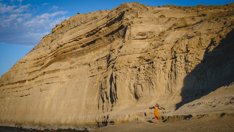 Audrey Bergner of That Backpacker stands at the base of dramatic sandstone cliffs during sunrise in Puerto Pirámides, Patagonia. Her small figure against the immense rock formations highlights the scale and rugged beauty of Peninsula Valdés’ coastal landscape