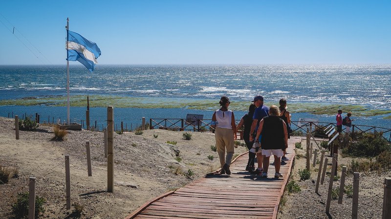 Visitors walking along a wooden boardwalk towards the coastline at Punta Norte on Península Valdés, Argentina, with the national flag waving in the wind. The viewpoint overlooks the Atlantic Ocean, a prime location for spotting sea lions and orcas in season