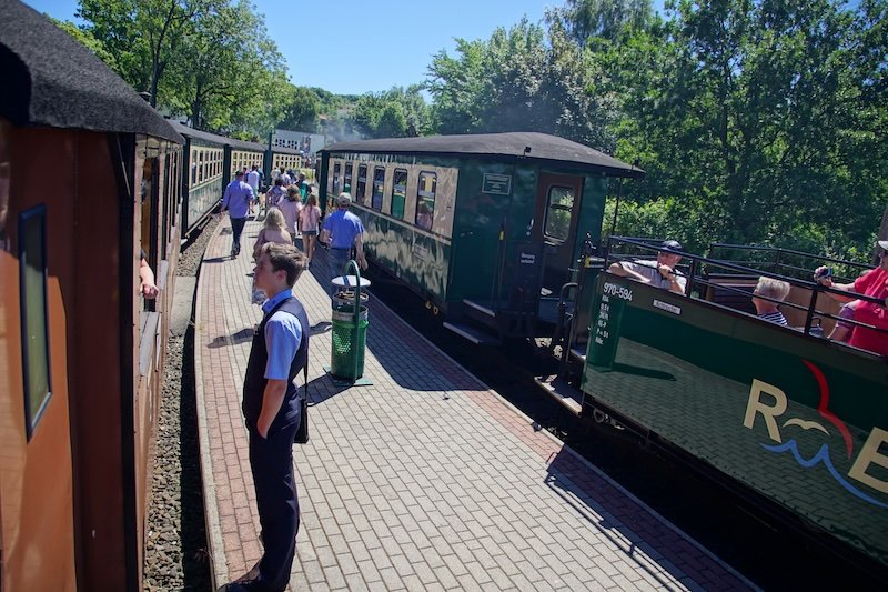 Passengers boarding the historic Rasender Roland narrow-gauge steam train at a station platform on Rügen Island, Germany, surrounded by lush summer greenery