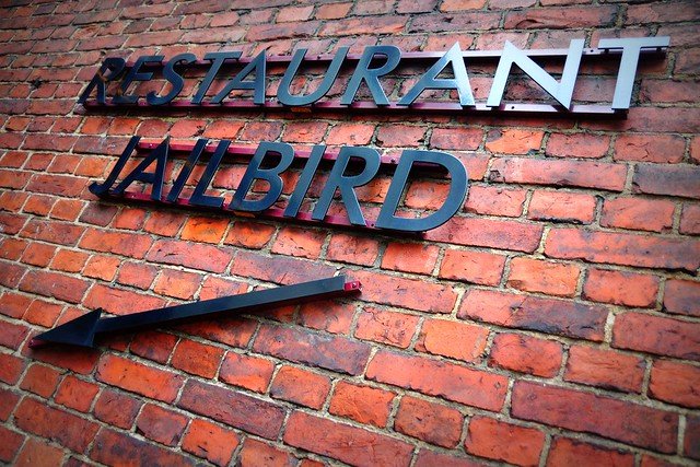 Restaurant Jailbird sign on a red-brick wall at Hotel Katajanokka in Helsinki, Finland, with an arrow pointing toward the entrance, highlighting the jailhouse-themed dining experience inside the historic prison hotel where guests enjoy breakfast