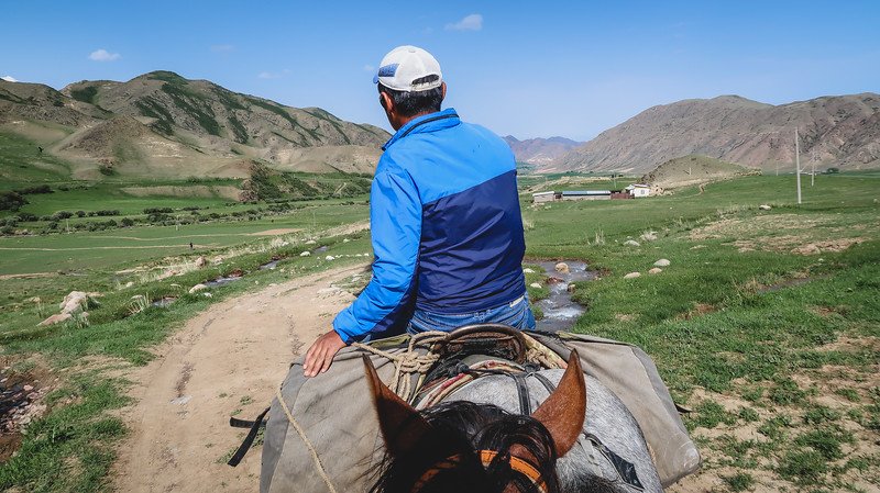 A horse rider (our guide) in a blue jacket and white cap heading towards a small rural town in Kyrgyzstan, riding along a dirt path through green valleys and rocky hills during the 3-Day Panorama Trek